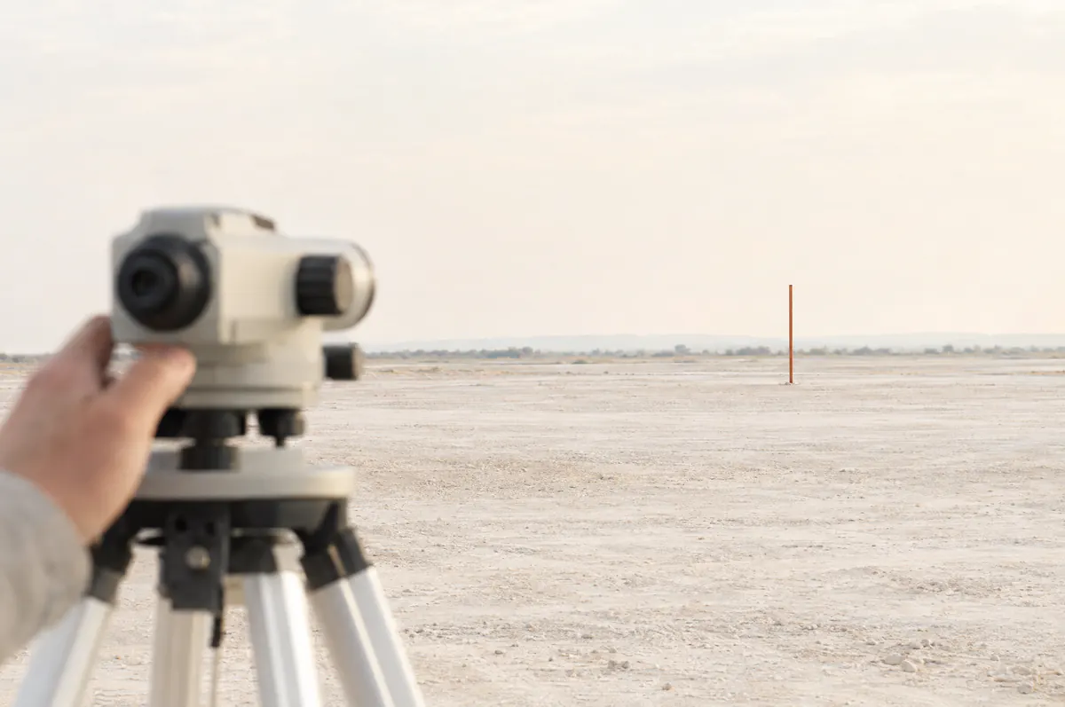 Illustration of a surveyor's level on a tripod, a hand on the instrument, aimed across a bright open site toward a distant rust-coloured ranging pole.